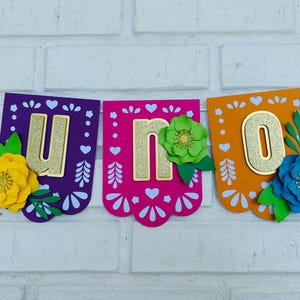 May include: A colorful paper banner with the word "UNO" in gold glitter letters. Each letter is on a separate pennant with a scalloped edge. The pennants are decorated with white hearts and flowers in yellow, green, and blue.