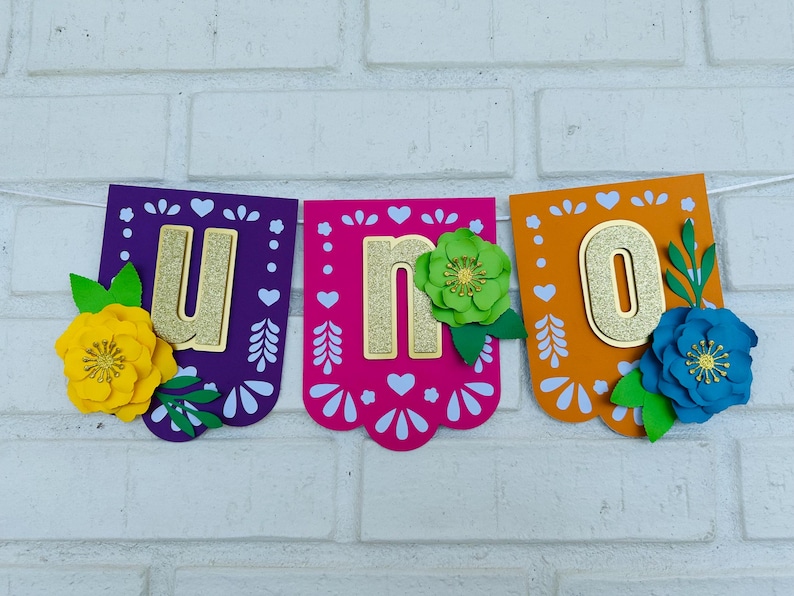 May include: A colorful paper banner with the word "UNO" in gold glitter letters. Each letter is on a separate pennant with a scalloped edge. The pennants are decorated with white hearts and flowers in yellow, green, and blue.