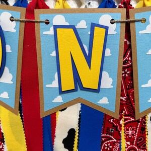 K&ouml;nnte beinhalten: Ein blaues und gelbes Banner mit dem Wort "ONE" in gro&szlig;en gelben Buchstaben. Das Banner ist mit wei&szlig;en Wolken auf blauem Hintergrund verziert. Das Banner ist an einer Schnur befestigt, an der rote, gelbe, blaue und wei&szlig;e Stoffstreifen h&auml;ngen.