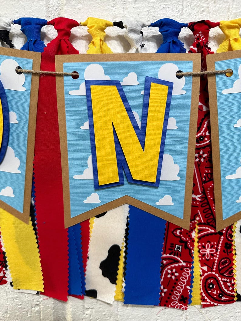 K&ouml;nnte beinhalten: Ein blaues und gelbes Banner mit dem Wort "ONE" in gro&szlig;en gelben Buchstaben. Das Banner ist mit wei&szlig;en Wolken auf blauem Hintergrund verziert. Das Banner ist an einer Schnur befestigt, an der rote, gelbe, blaue und wei&szlig;e Stoffstreifen h&auml;ngen.