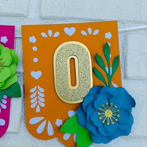 May include: Orange and white paper banner with a gold glitter number "0" and blue and green paper flowers. The banner has a traditional Mexican design.