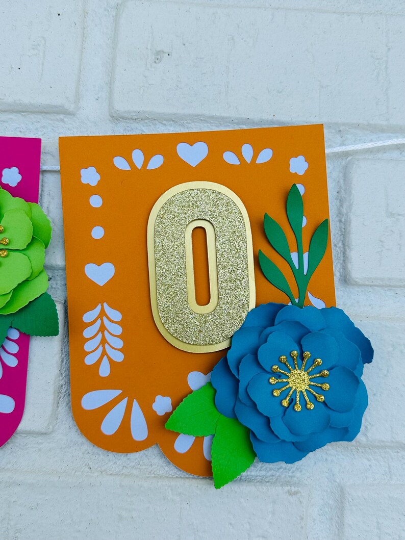 May include: Orange and white paper banner with a gold glitter number "0" and blue and green paper flowers. The banner has a traditional Mexican design.