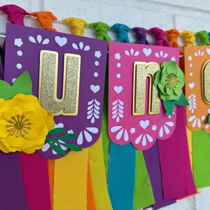 May include: A colorful "Uno" banner with gold glitter letters and paper flowers. The banner is made of purple, pink, orange, and green paper and has a rainbow fringe.