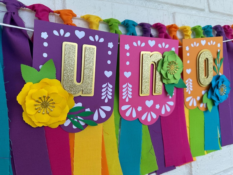 May include: A colorful "Uno" banner with gold glitter letters and paper flowers. The banner is made of purple, pink, orange, and green paper and has a rainbow fringe.