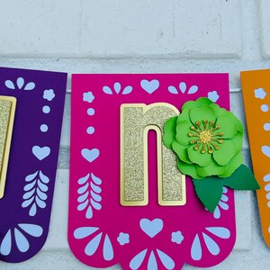 May include: A pink, purple, and orange paper banner with the letter "n" in gold glitter. The letter is decorated with a green paper flower and leaves.