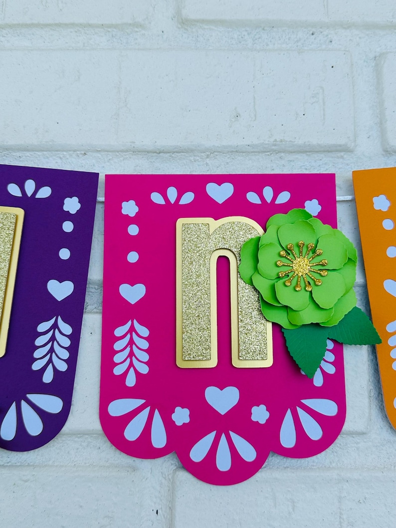 May include: A pink, purple, and orange paper banner with the letter "n" in gold glitter. The letter is decorated with a green paper flower and leaves.