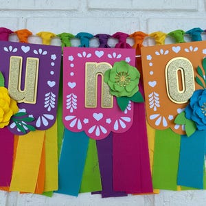 May include: A colorful paper banner with the word "UNO" in gold glitter letters. Each letter is on a separate card with a white scalloped edge and a colorful floral design. The banner is strung with rainbow colored fabric streamers.
