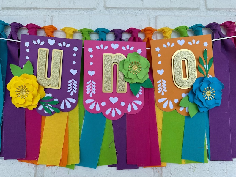 May include: A colorful paper banner with the word "UNO" in gold glitter letters. Each letter is on a separate card with a white scalloped edge and a colorful floral design. The banner is strung with rainbow colored fabric streamers.