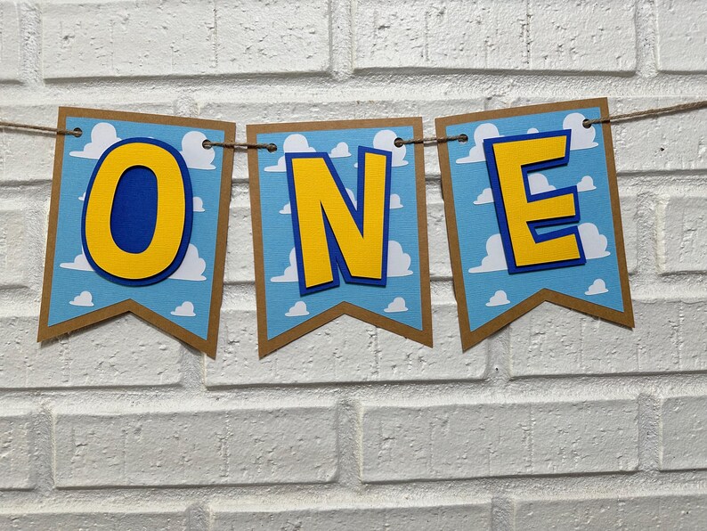 K&ouml;nnte beinhalten: Ein Banner mit dem Wort "ONE" in gelben Buchstaben mit blauen Umrandungen auf blau-wei&szlig;em Hintergrund. Das Banner ist aus Papier und hat einen braunen Rand.