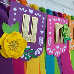 May include: A colorful banner with the word "UNO" in gold glitter letters. The banner is decorated with paper flowers and has rainbow-colored fabric streamers hanging from the bottom.