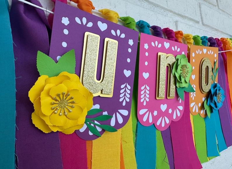 May include: A colorful banner with the word "UNO" in gold glitter letters. The banner is decorated with paper flowers and has rainbow-colored fabric streamers hanging from the bottom.