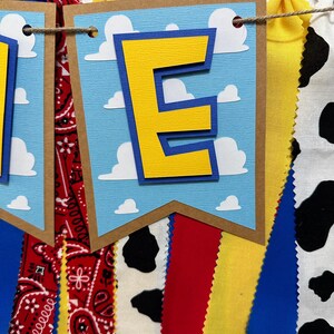 K&ouml;nnte beinhalten: Ein blaues und gelbes Banner mit dem Buchstaben "E" in der Mitte. Das Banner ist mit wei&szlig;en Wolken verziert und hat einen braunen Rand. Das Banner ist an einer Schnur mit roten, gelben, blauen und schwarz-wei&szlig;en Stoffstreifen befestigt.