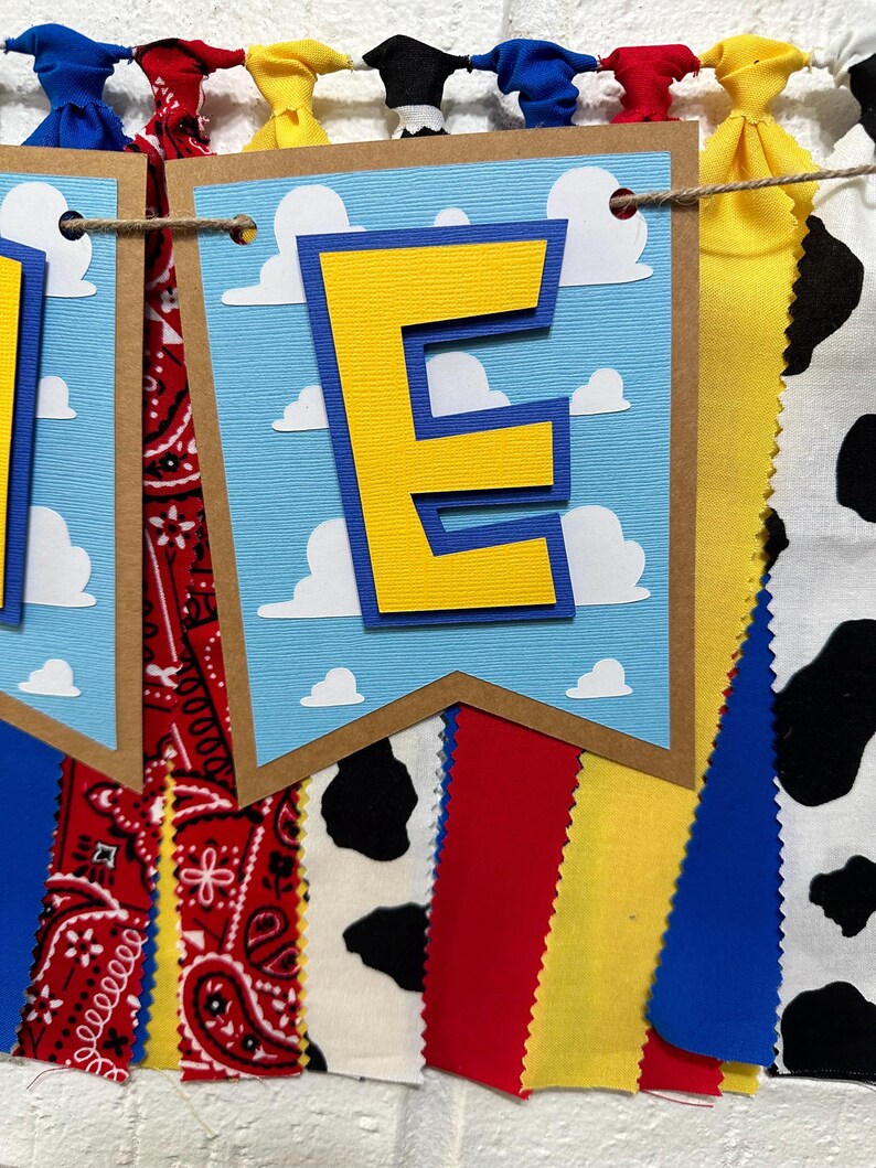 K&ouml;nnte beinhalten: Ein blaues und gelbes Banner mit dem Buchstaben "E" in der Mitte. Das Banner ist mit wei&szlig;en Wolken verziert und hat einen braunen Rand. Das Banner ist an einer Schnur mit roten, gelben, blauen und schwarz-wei&szlig;en Stoffstreifen befestigt.
