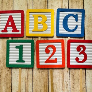 May include: Six colorful cardboard blocks with letters A, B, C and numbers 1, 2, 3. The blocks are on wooden sticks and are arranged in a row.