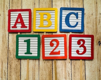 Könnte beinhalten: Sechs bunte Pappkartons mit den Buchstaben A, B, C und den Zahlen 1, 2, 3. Die Kartons sind auf Holzstäbchen und in einer Reihe angeordnet.