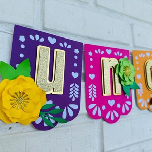 May include: A colorful paper banner with the word "UNO" in gold glitter letters. Each letter is on a separate piece of paper with a different color and design. The letters are decorated with paper flowers.
