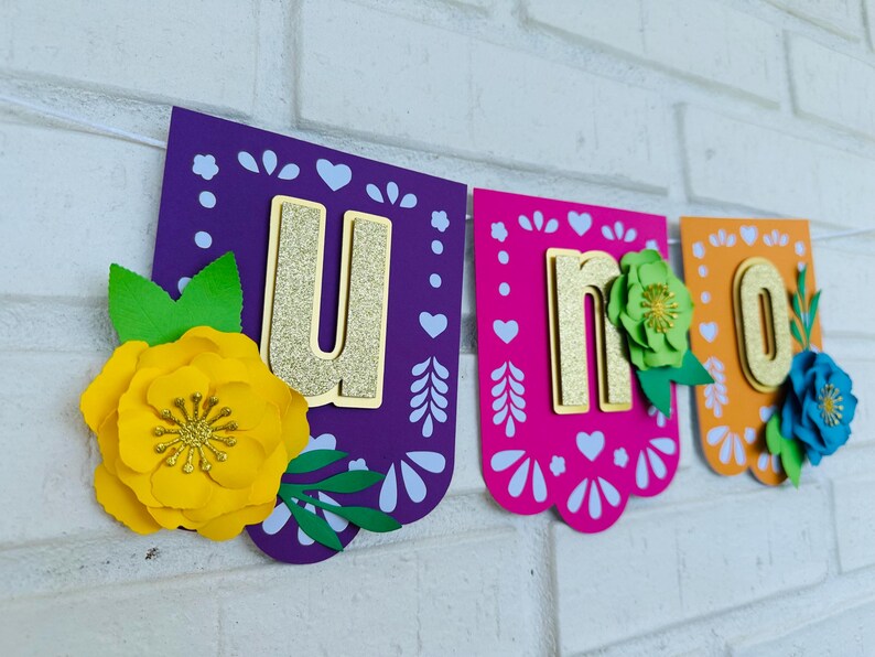 May include: A colorful paper banner with the word "UNO" in gold glitter letters. Each letter is on a separate piece of paper with a different color and design. The letters are decorated with paper flowers.
