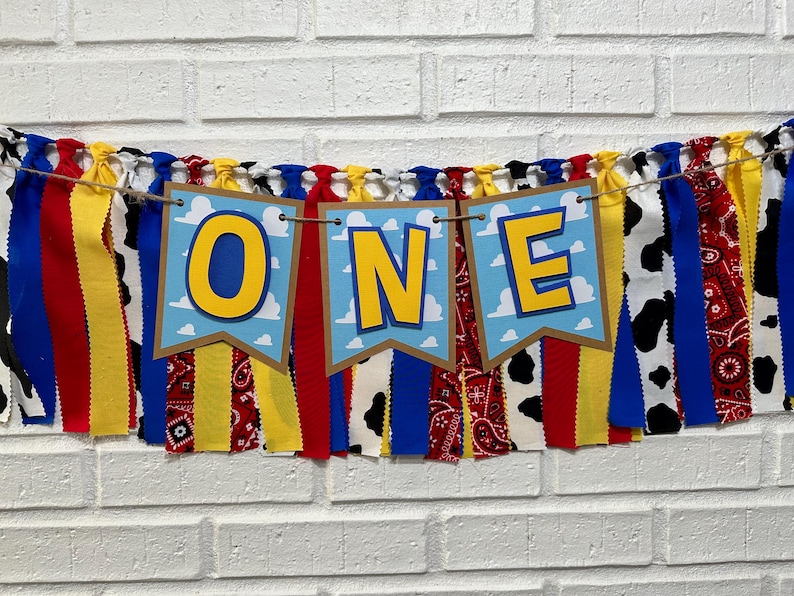 K&ouml;nnte beinhalten: Ein farbenfrohes Stoffbanner mit dem Wort "ONE" in gelben Buchstaben auf blauem Hintergrund. Das Banner ist mit roten, gelben, blauen und wei&szlig;en Stoffstreifen verziert. Das Banner ist perfekt f&uuml;r eine erste Geburtstagsfeier.