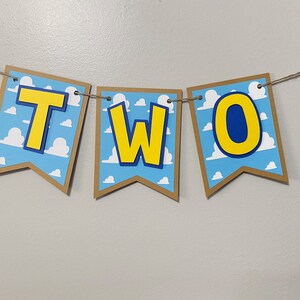 K&ouml;nnte beinhalten: Ein braunes und gelbes Banner mit dem Wort "ZWEI" in gelben Buchstaben mit blauen Umrandungen. Das Banner hat einen blauen und wei&szlig;en Hintergrund mit einem Wolkenmuster.