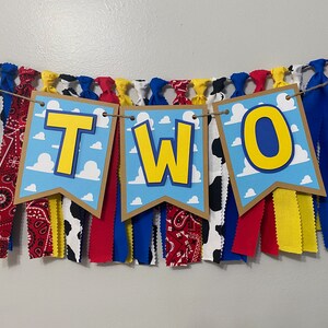 K&ouml;nnte beinhalten: Ein Banner mit dem Wort "ZWEI" in gelben Buchstaben auf blauem Hintergrund mit wei&szlig;en Wolken. Das Banner ist aus braunem Papier und hat einen Fransen aus rotem, gelbem, blauem und wei&szlig;em Stoff.