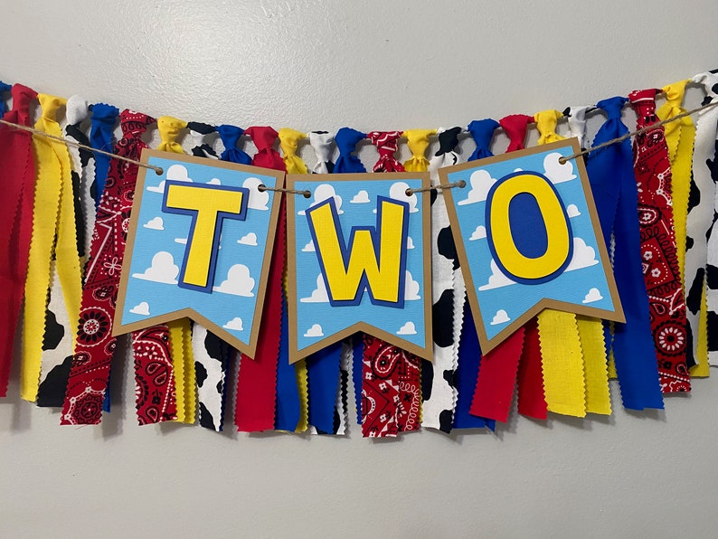 K&ouml;nnte beinhalten: Ein Banner mit dem Wort "ZWEI" in gelben Buchstaben auf blauem Hintergrund mit wei&szlig;en Wolken. Das Banner ist aus braunem Papier und hat einen Fransen aus rotem, gelbem, blauem und wei&szlig;em Stoff.