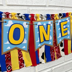 K&ouml;nnte beinhalten: Ein farbenfrohes Banner mit dem Wort "ONE" in gelben Buchstaben auf blauem Hintergrund mit wei&szlig;en Wolken. Das Banner besteht aus Stoffresten in verschiedenen Mustern, darunter rotes und wei&szlig;es Gingham, schwarz-wei&szlig;es Kuhmuster und blau-wei&szlig;es Paisley.