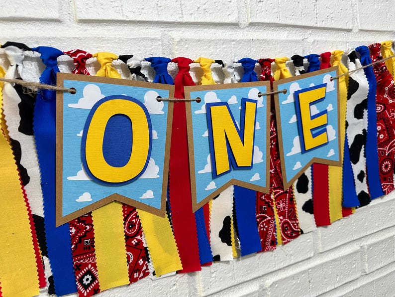 K&ouml;nnte beinhalten: Ein farbenfrohes Banner mit dem Wort "ONE" in gelben Buchstaben auf blauem Hintergrund mit wei&szlig;en Wolken. Das Banner besteht aus Stoffresten in verschiedenen Mustern, darunter rotes und wei&szlig;es Gingham, schwarz-wei&szlig;es Kuhmuster und blau-wei&szlig;es Paisley.