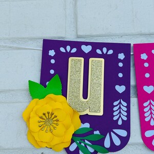 May include: Purple and pink paper banner with a gold glitter letter "U" and a yellow paper flower. The banner has a white cut-out design.