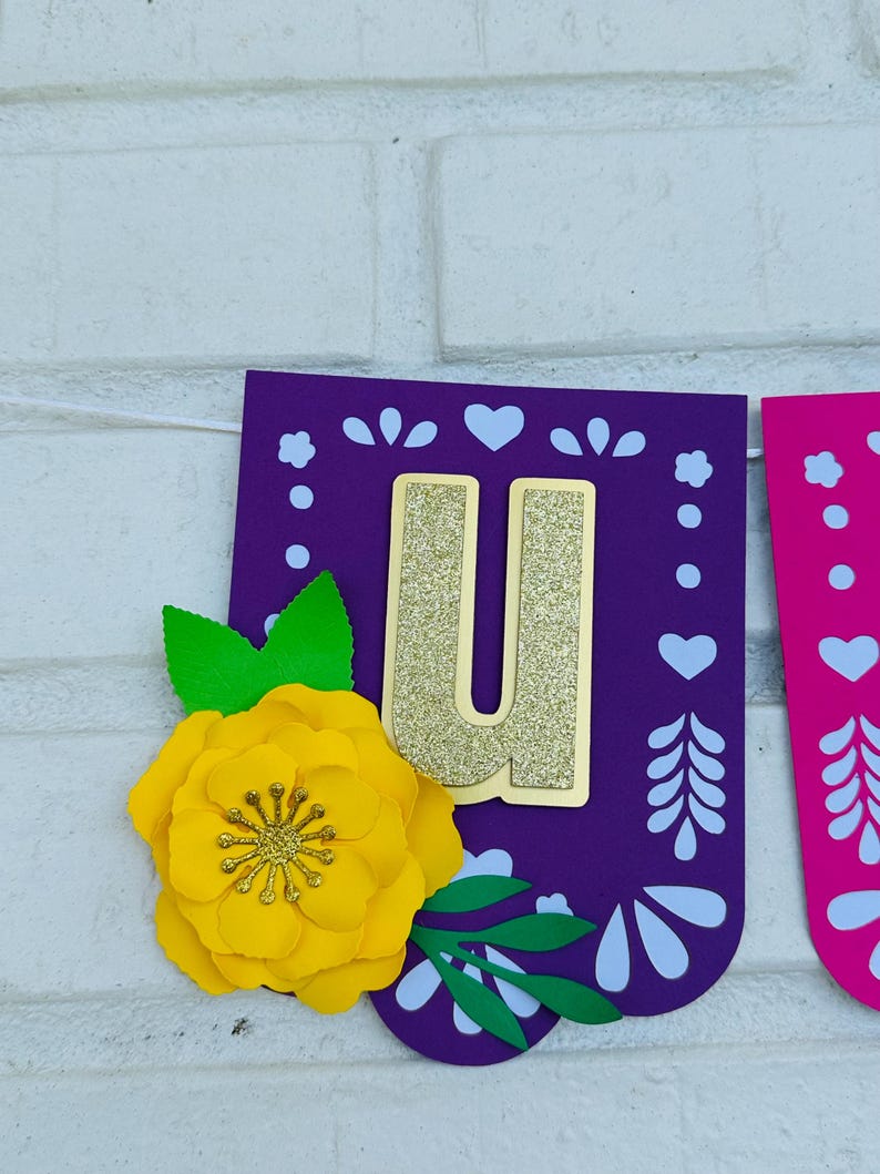 May include: Purple and pink paper banner with a gold glitter letter "U" and a yellow paper flower. The banner has a white cut-out design.