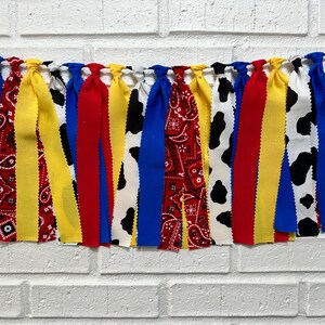 K&ouml;nnte beinhalten: Ein farbenfrohes Stoffbanner mit roten, gelben, blauen und wei&szlig;en Streifen. Das Banner zeigt ein Kuhmuster und ein rotes Bandanamuster.