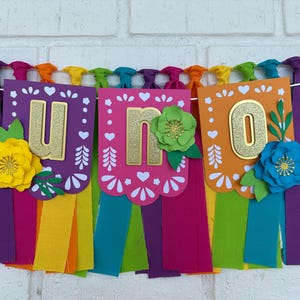 May include: A colorful banner with the word "UNO" in gold glitter letters. The banner is decorated with paper flowers and has colorful tassels hanging from the bottom.