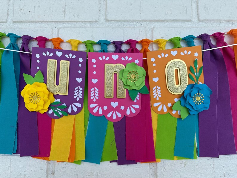 May include: A colorful banner with the word "UNO" in gold glitter letters. The banner is decorated with paper flowers and has colorful tassels hanging from the bottom.