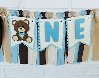 May include: A blue and brown banner with the word "ONE" and a brown teddy bear wearing a blue bow tie. The banner is made of felt and has a scalloped edge. It is decorated with blue, brown, and beige ribbons.