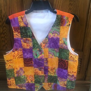 Puede incluir: Un chaleco patchwork con diseño de cuello en V, con una vibrante mezcla de patrones florales naranjas, morados y verdes. El chaleco tiene un forro blanco y detalles naranjas en los hombros. Se muestra en una percha negra.