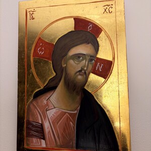 May include: A religious icon featuring a depiction of Jesus Christ. The artwork has a gold background and a halo with a red cross. Jesus is depicted with brown hair and a beard, wearing a pink robe and black cloak. The icon has text in the upper corners.