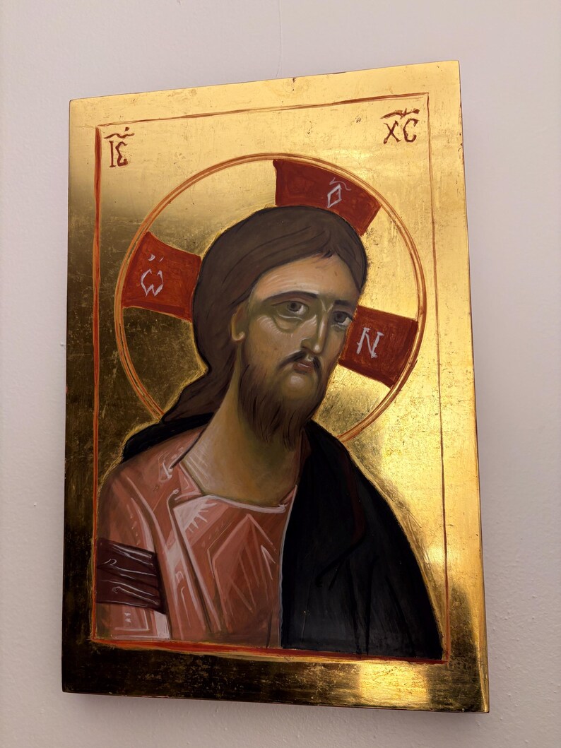 May include: A religious icon featuring a depiction of Jesus Christ. The artwork has a gold background and a halo with a red cross. Jesus is depicted with brown hair and a beard, wearing a pink robe and black cloak. The icon has text in the upper corners.