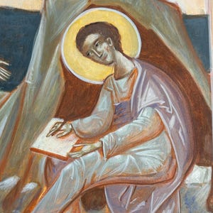 May include: An icon painting depicting a figure with a golden halo, dressed in white and lavender robes, seated and writing on a parchment. The artwork features warm tones of brown, orange, and beige, with a focus on the figure's contemplative expression.