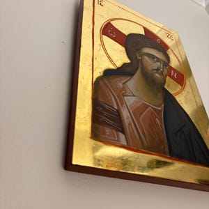 May include: Icon of a figure with long brown hair and a beard, wearing sunglasses. The figure is depicted in a red and gold halo, with a gold background and a dark brown frame. The image is mounted on a wall.