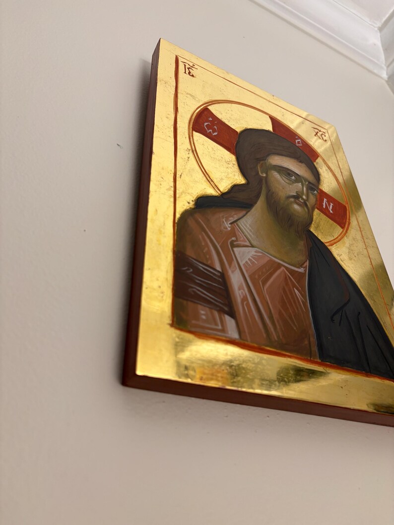 May include: Icon of a figure with long brown hair and a beard, wearing sunglasses. The figure is depicted in a red and gold halo, with a gold background and a dark brown frame. The image is mounted on a wall.