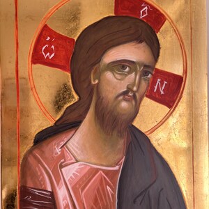 May include: An icon depicting a figure with brown hair and a beard, wearing a pink robe and a dark cloak. A red halo with a cross shape surrounds the figure's head. The background is gold.