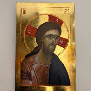 May include: A religious icon featuring a portrait of Jesus Christ. The artwork depicts Christ with brown hair and a beard, wearing a red and black robe. The background is gold with red accents and Greek letters.