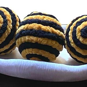 May include: Three black and gold striped balls in a white bowl. The balls are made of a soft, fuzzy material.