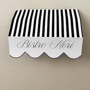 Peut inclure: Panneau mural décoratif avec un motif d'auvent à rayures noires et blanches. Le panneau porte l'inscription "Bistro Nori" en écriture cursive. En dessous, deux gamelles noires pour animaux de compagnie sur un support en bois.