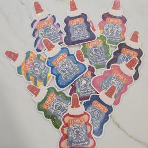 May include: A collection of holographic stickers shaped like glue bottles, each featuring a unique character illustration and the text UMA'S followed by a specific edition name. The stickers display various colors including red, blue, green, and purple, with a shimmering finish that reflects light across the surface of each individual adhesive design.