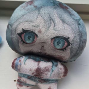 May include: A plush toy featuring a stylized character head with large blue eyes, pale skin, and blue hair, covered in dark red splatters. The small, rounded body also displays the same red splatter pattern, with a white loop attached to the top.