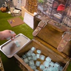 May include: A wooden chest filled with numerous small, clear plastic containers, each holding a light blue square item. A silver metal tray sits beside the chest, and a hand is placing another small container onto the tray. A sign reads Wildwood Scoop.