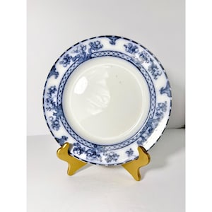 May include: White plate with a blue floral and geometric border design. The plate is displayed on a gold-colored stand. The plate's rim features a detailed pattern of flowers and foliage.
