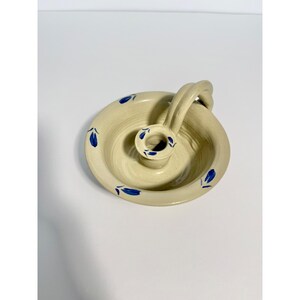 May include: Cream-colored ceramic candle holder with a circular base and a curved handle. The holder features a central well for the candle and is decorated with blue floral accents. The handle is arched, providing a comfortable grip.