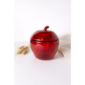 May include: A red glass apple-shaped jar with a lid and a stem. The jar is sitting on a white surface with wheat stalks and a beige cloth in the background. The jar is a decorative item.
