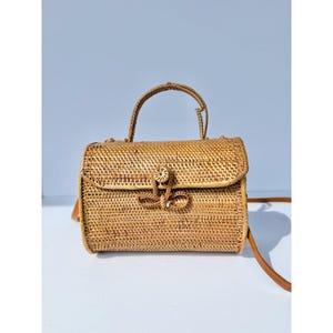 May include: A cylindrical woven rattan handbag featuring a top handle and a long shoulder strap. The bag has a flap closure secured by a circular rattan button and a decorative woven loop. The natural light brown material has a textured finish.
